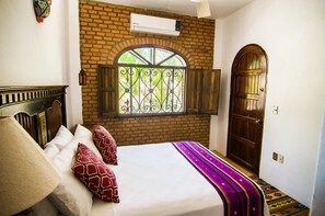 Una camera d'albergo con parete in mattoni, finestra con inferriate decorative, porta in legno e letto con cuscini a motivi.