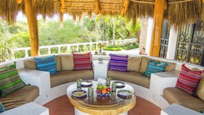 A covered outdoor seating area with a round table, colorful cushions, and a view of lush greenery.