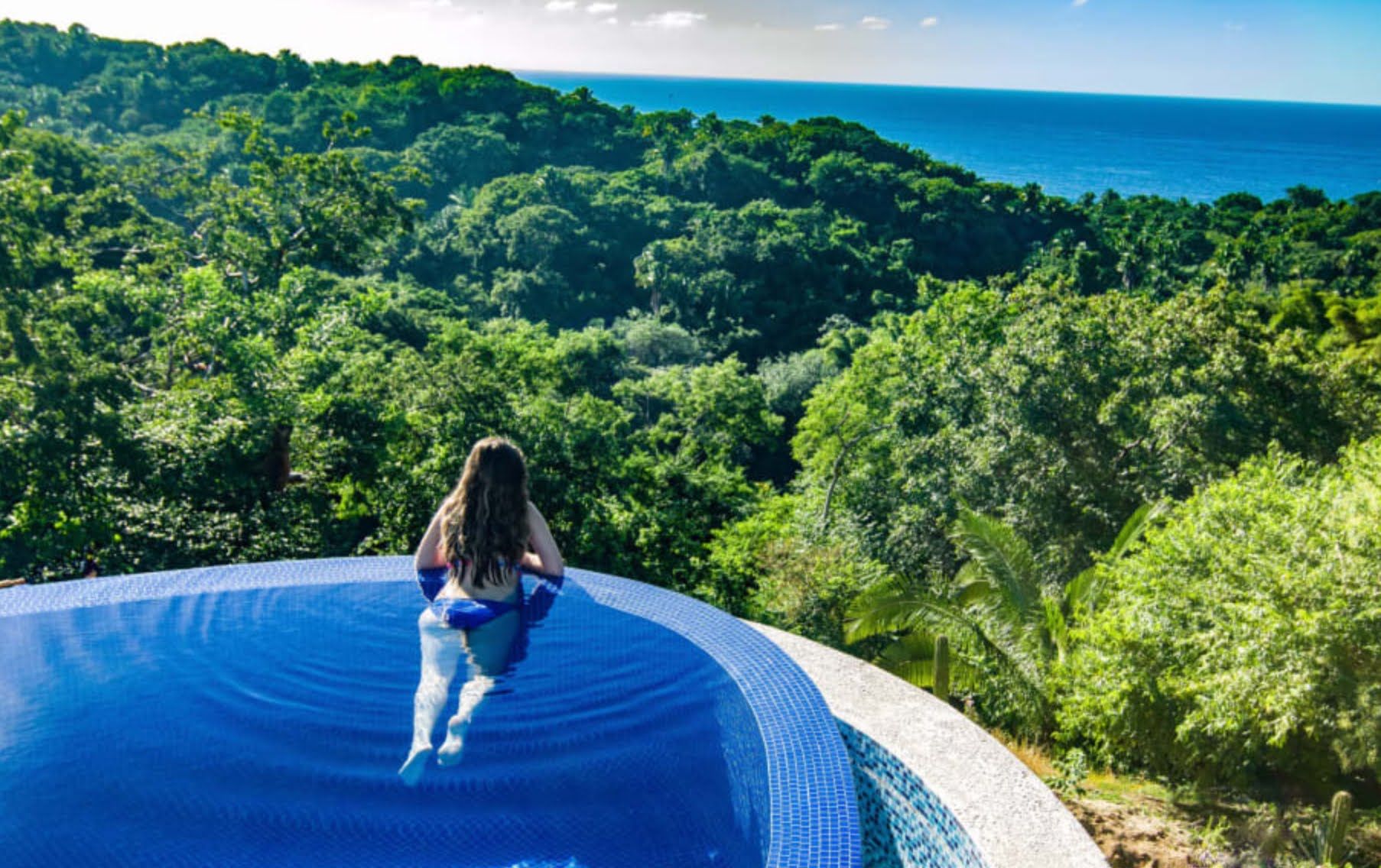 Eine Person sitzt am Poolrand mit Blick auf einen Wald und den Ozean.