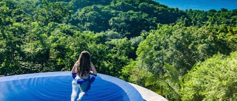 A person sitting on the edge of a pool with a view of a forest and the ocean.