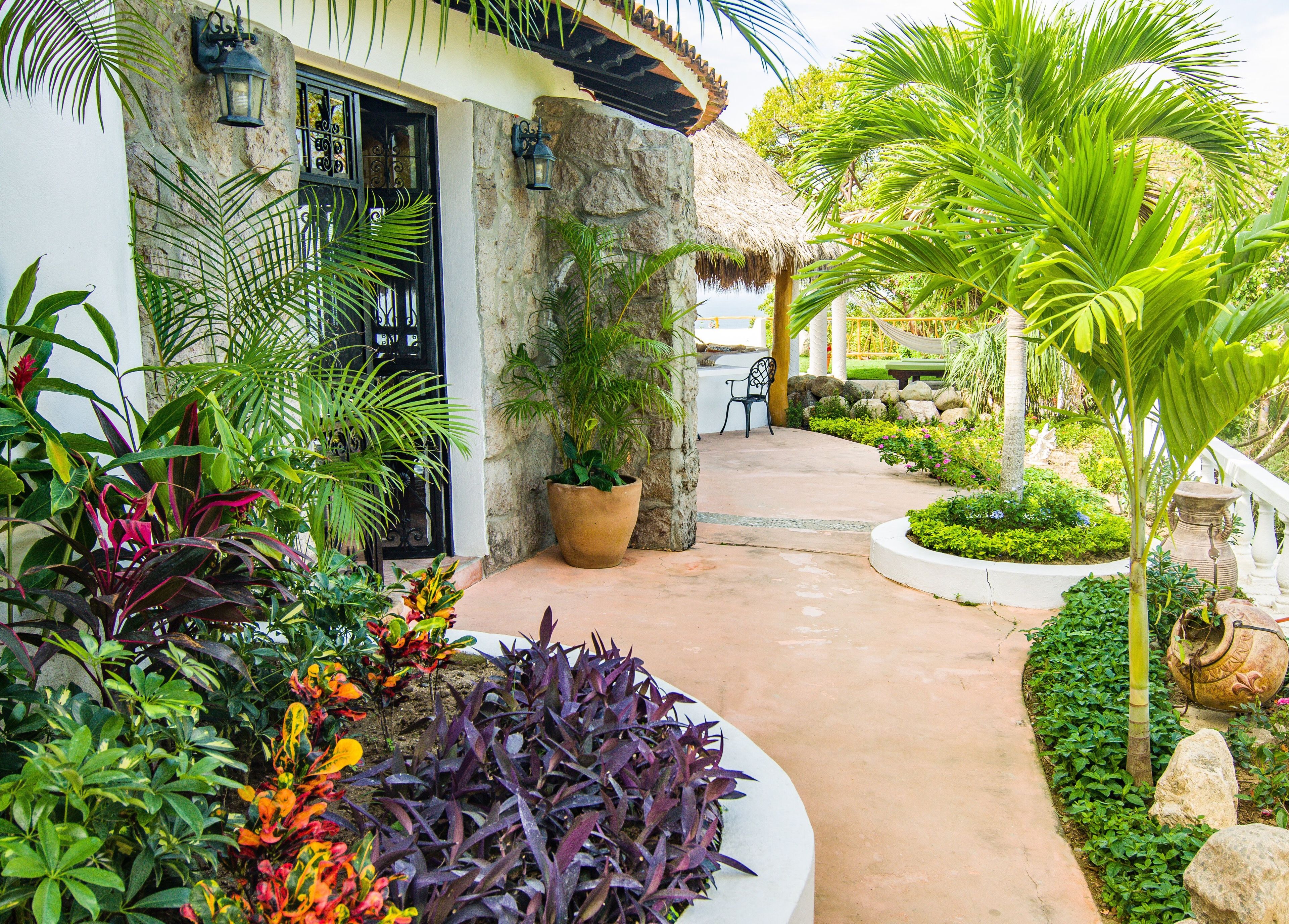 Un sendero en un jardín tropical que conduce a un edificio con fachada de piedra y techo de paja.