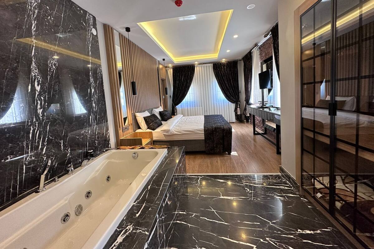 A modern hotel room with a large marble bathtub, a neatly made bed, a small bedside table, and a large mirror.