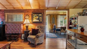 A cozy living room with a patterned armchair, a wooden ceiling, and a dining area with a table and chairs. - Walk Softly, There’s a Dream Here! (Colchester)