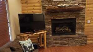 A living room with a stone fireplace, a flat-screen TV on a wooden stand, a brown sofa with patterned cushions, and a wooden coffee table.