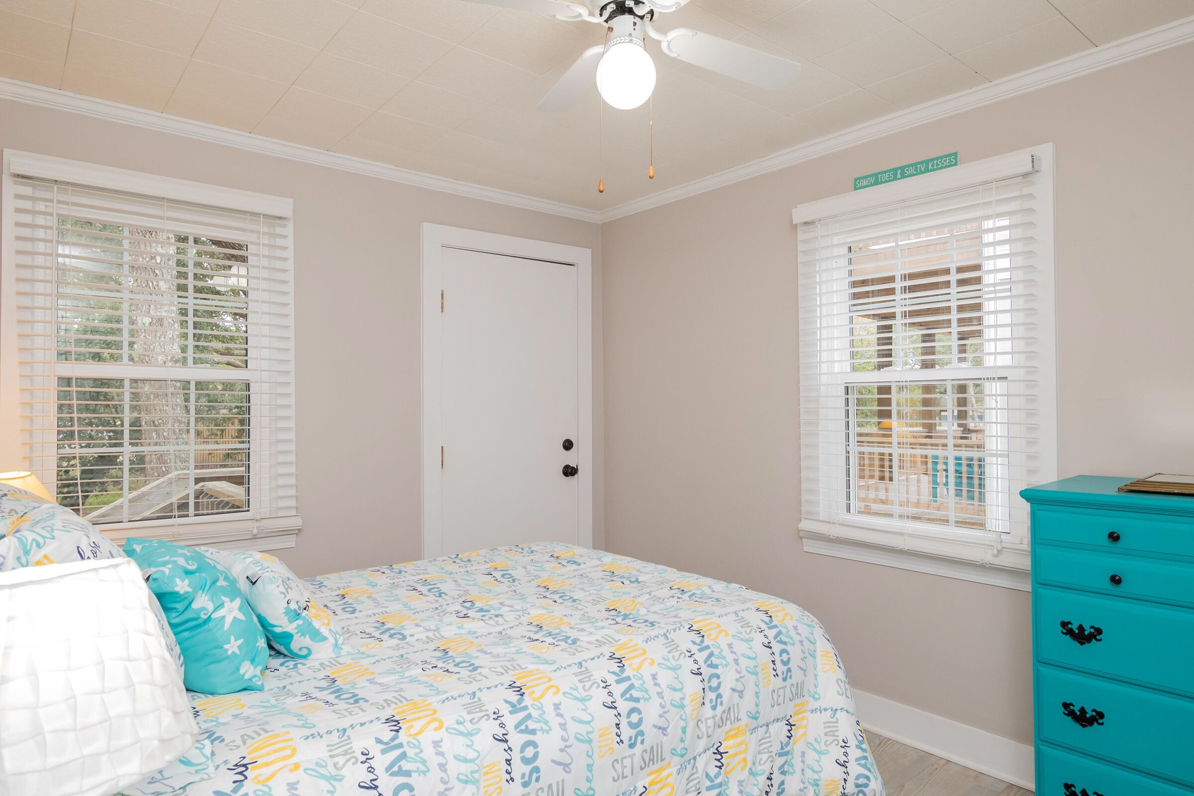 Een slaapkamer met een bed, twee ramen met zonwering, een plafondventilator en een turquoise dressoir.