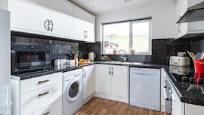 A modern kitchen with white cabinets, a black countertop, a built-in washing machine, and a window providing natural light. - Coastal eco-home for up to 6, short walk from Porthtowan village and beach (Porthtowan)