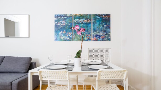 Ein Essbereich mit einem weißen Tisch, gedeckt für vier Personen, umgeben von weißen Stühlen, einem grauen Sofa und einem großen Blumenbild an der Wand.