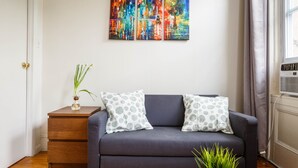 Ein Wohnzimmer mit einem dunkelblauen Sofa, zwei gemusterten Kissen, einem kleinen Holztisch mit einer Vase und einer Topfpflanze.