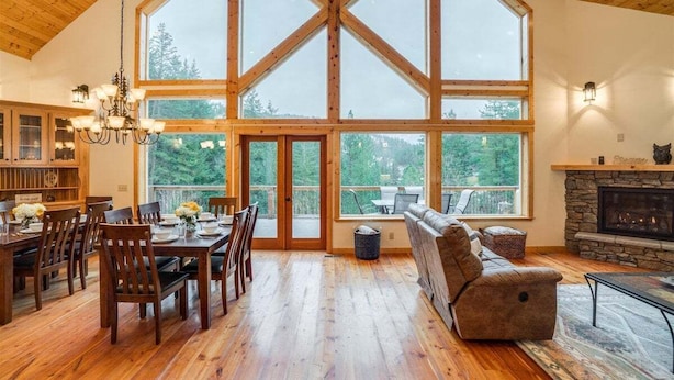A spacious room with a high ceiling, wooden beams, a dining area with a chandelier, a fireplace, and large windows offering a view of the outdoors.