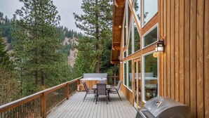 A wooden deck with a hot tub, grill, and dining area overlooking a forested hillside.