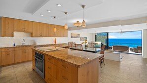 A modern kitchen with wooden cabinets, granite countertops, and a large island. It features a dining area with wicker furniture and a view of the ocean. - Sapphire Lotus~Ocean Front~A/C~Pool~BBQ (Princeville)