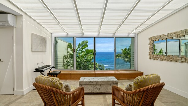 A room with a large sliding glass door leading to a balcony with a view of the ocean. There are two wicker chairs and a small table with a keyboard. - Sapphire Lotus~Ocean Front~A/C~Pool~BBQ (Princeville)