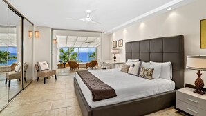 A modern bedroom with a large bed, a ceiling fan, a nightstand, and a view of the ocean through large windows. - Sapphire Lotus~Ocean Front~A/C~Pool~BBQ (Princeville)