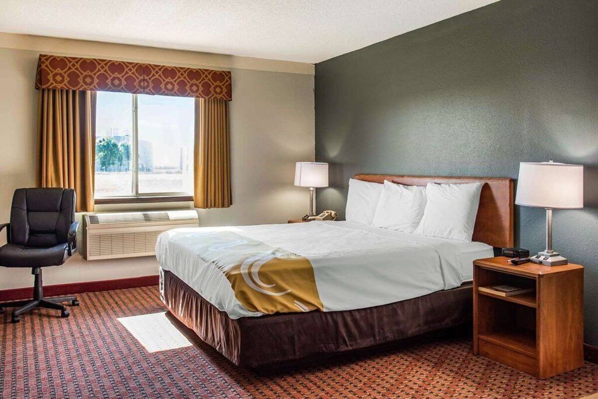 A hotel room with a bed, two bedside lamps, a desk with a lamp, a chair, and a window with curtains.