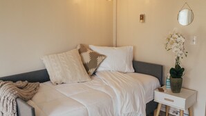 Una cama bien hecha con ropa blanca y almohadas estampadas, una mesita de noche con una planta y un espejo montado en la pared.