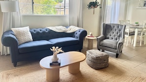 Una sala de estar con un sofá azul, un sillón gris, una mesa de centro de madera y una pequeña mesa redonda con un jarrón y libros.