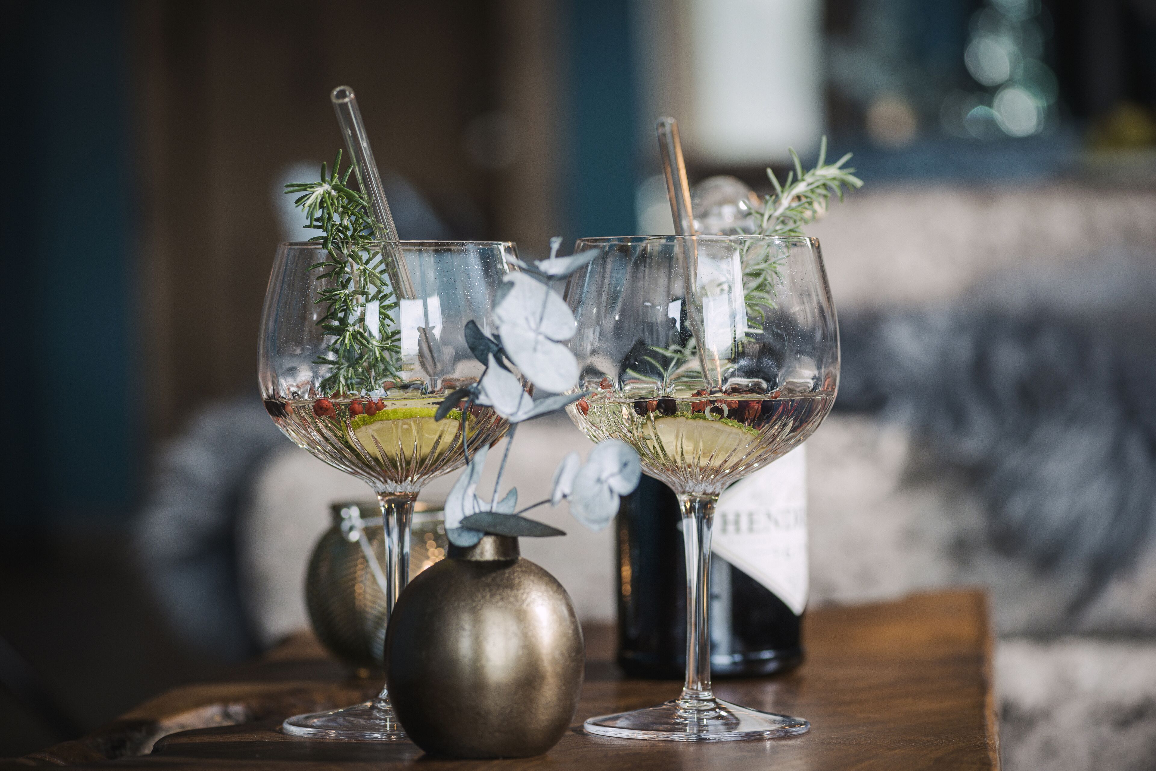 Two wine glasses with garnishes on a wooden table, a decorative vase, and a bottle in the background.