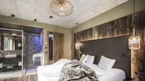 A modern hotel room with a large bed, a glass-enclosed shower, a wooden accent wall, and a minimalist design.