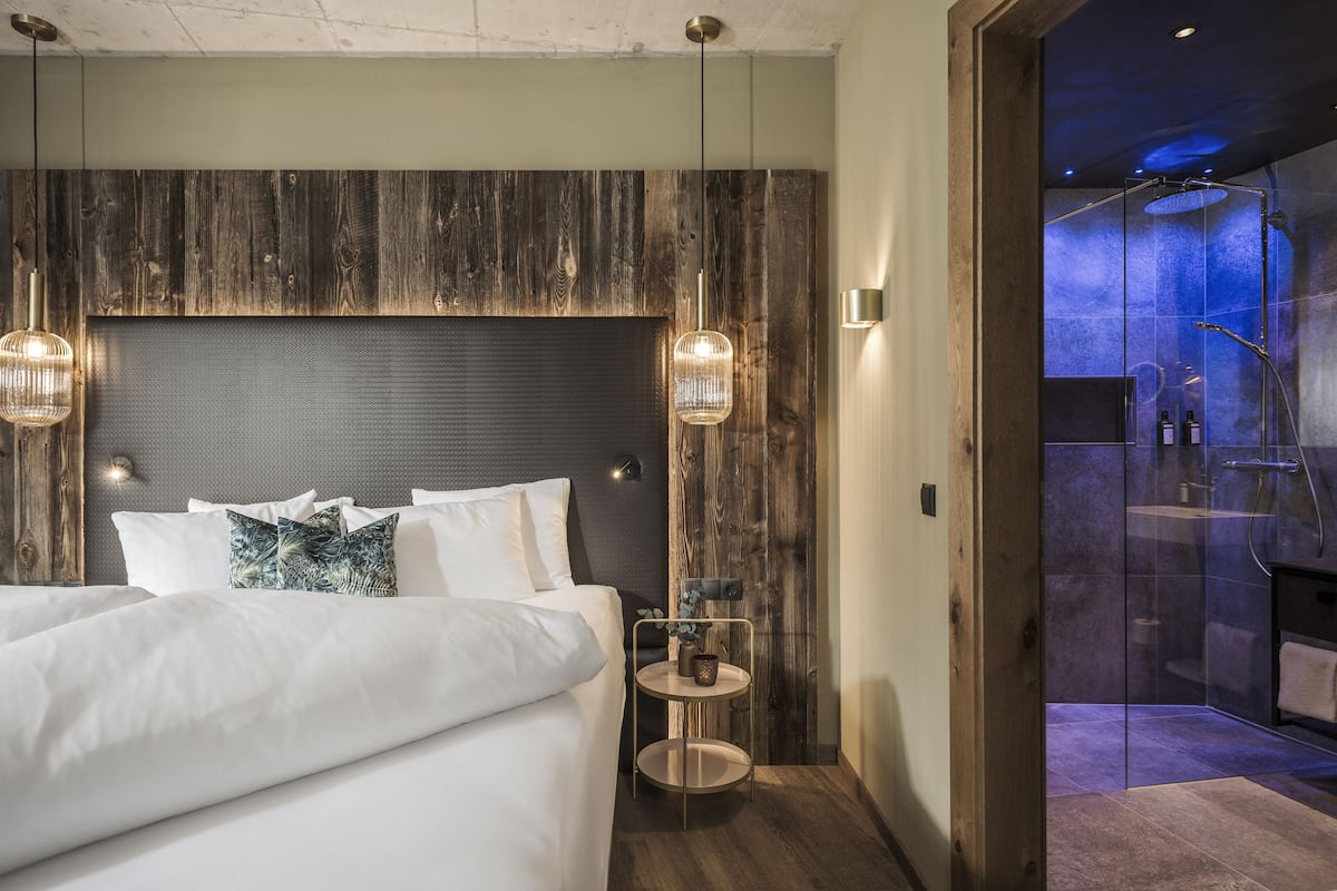 Una habitación de hotel moderna con una cama grande, cabecera de madera, dos lámparas colgantes, una pequeña mesa redonda con un florero y una zona de ducha con paredes de cristal.