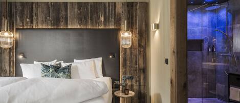 A modern hotel room with a large bed, wooden headboard, two pendant lights, a small round table with a vase, and a glass-enclosed shower area.