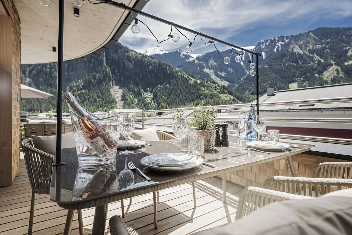 Mesa de comedor preparada con platos, vasos y una botella de vino en una terraza con vistas a la montaña.
