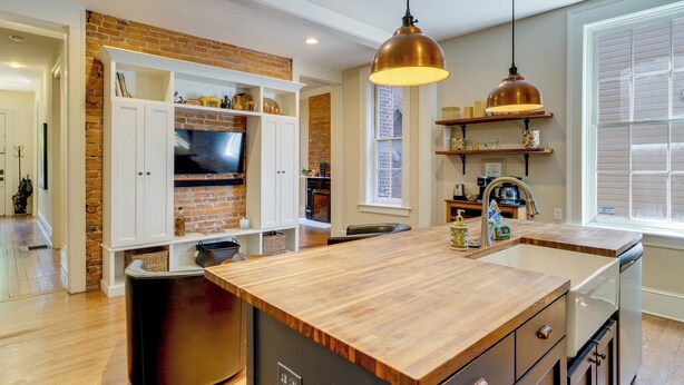 A kitchen with a wooden island, white cabinets, a brick accent wall, and a hanging pendant light.