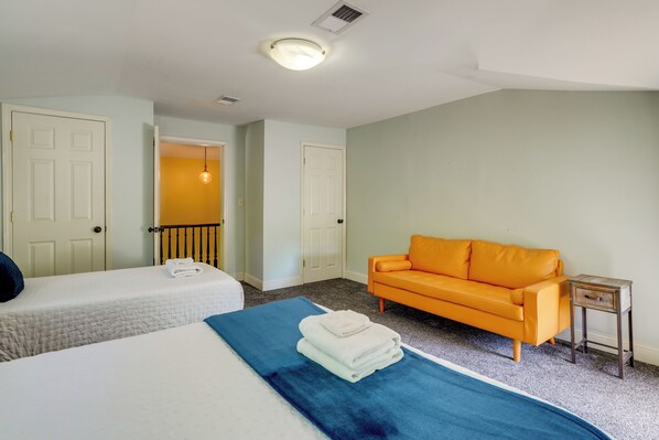 Habitación de hotel con dos camas, un sofá naranja, una mesita de madera y un pasillo que conduce a otra habitación.