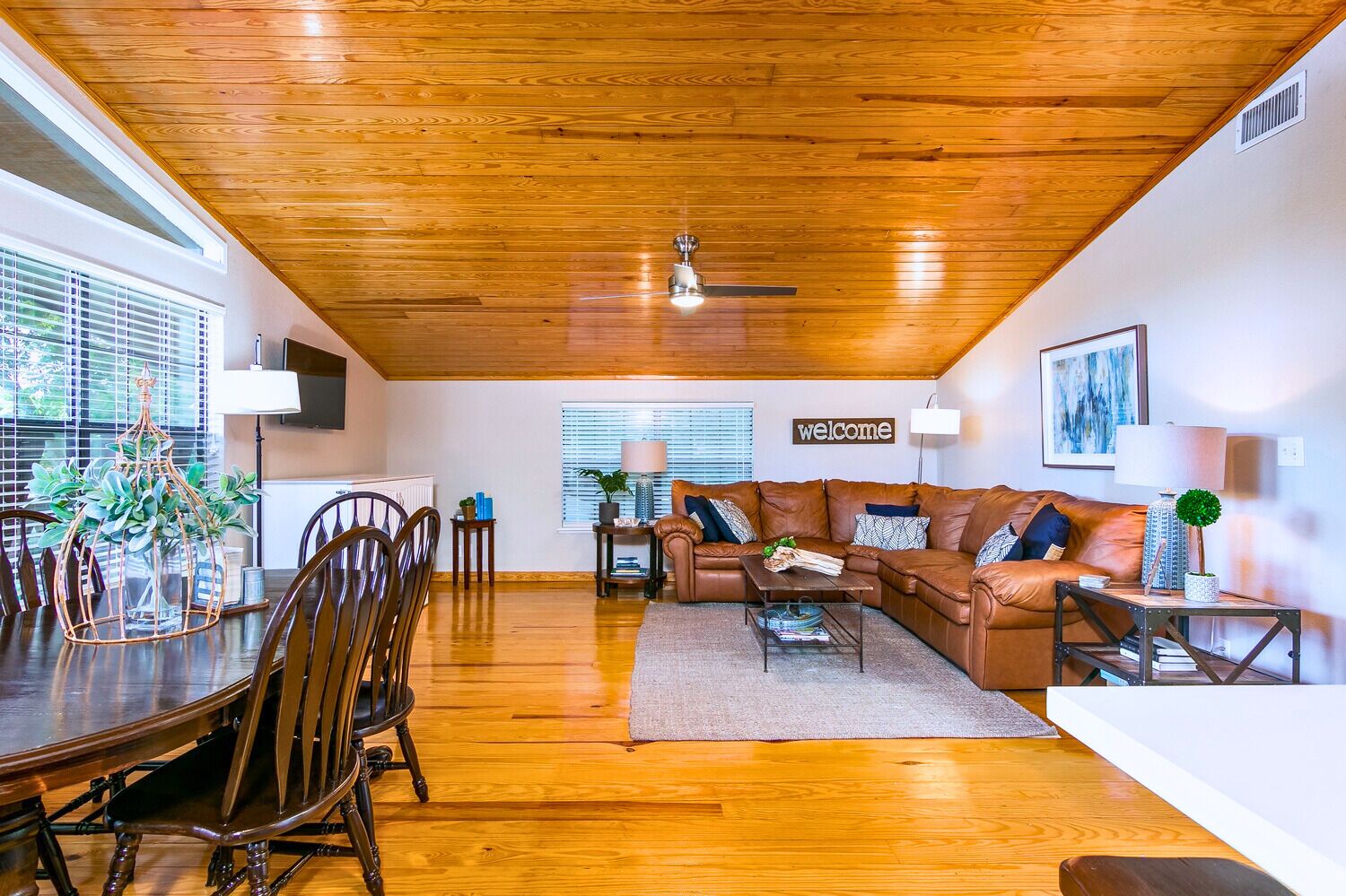 A spacious living room with a wooden ceiling, a brown leather sofa, a dining table with chairs, and a 'welcome' sign on the wall.
