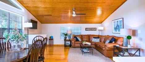 A spacious living room with a wooden ceiling, a brown leather sofa, a dining table with chairs, and a 'welcome' sign on the wall.