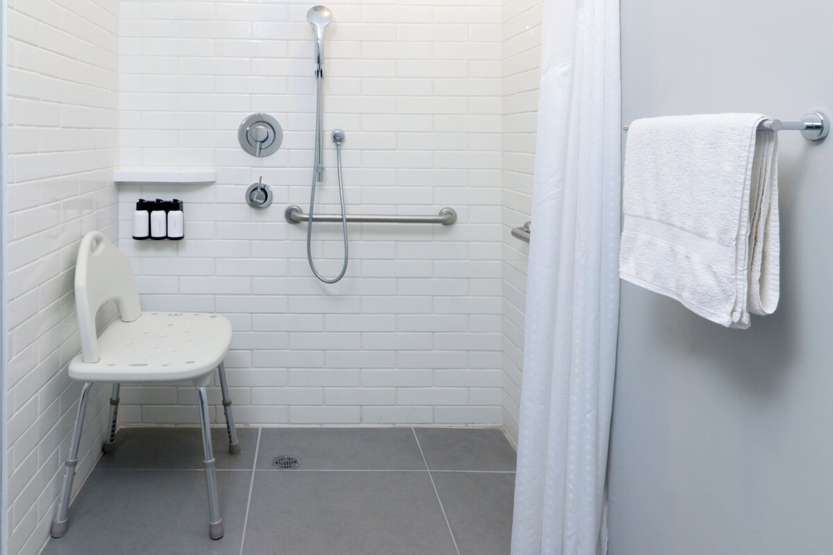 A bathroom with white tiles, a shower with a handheld showerhead, a grab bar, a white chair, and a towel rack.