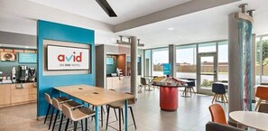 A modern hotel lobby with a central dining area, a reception desk, and a large window offering a view of the outdoors.