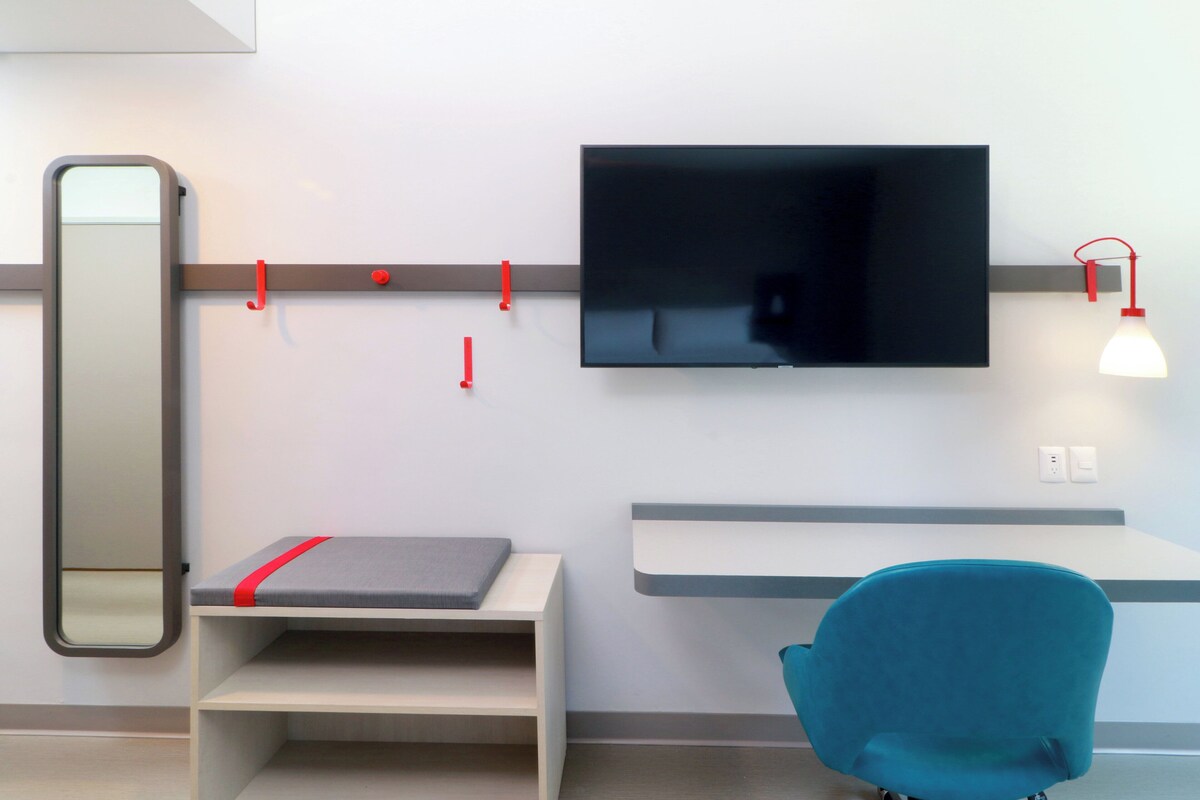 A modern room with a flat-screen TV mounted on the wall, a small desk with a chair, and a mirror on the left.