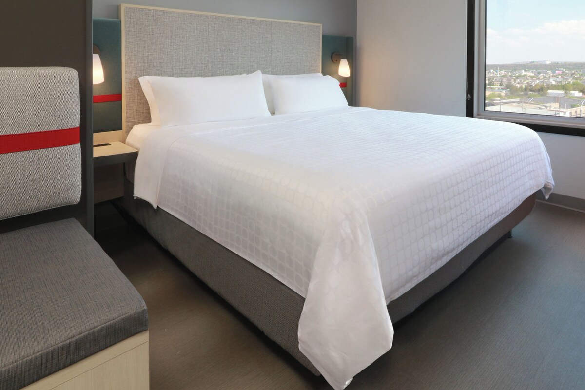 A neatly made bed with white bedding, a grey headboard, and a view of a cityscape through a window.
