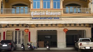 Un immeuble à plusieurs étages doté de balcons, une enseigne indiquant « Kampot Moyura Guest House », et plusieurs véhicules stationnés.