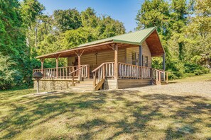 A wooden cabin with a porch, stone foundation, and a green roof, surrounded by trees. - Rustic Cabin w/ Hot Tub Near Broken Bow Lake! (Broken Bow)