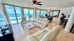 A spacious living room with a large window overlooking the ocean, a ceiling fan, and a dining area with a view. - Shores Club Direct Oceanfront 3/3 on Traffic Free Beach - Unit 901 (Daytona Beach Shores)