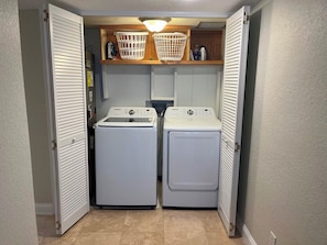 Une buanderie équipée de deux machines à laver et deux sèche-linge à chargement frontal, tous deux de couleur blanche, avec des étagères en bois au-dessus et un luminaire.