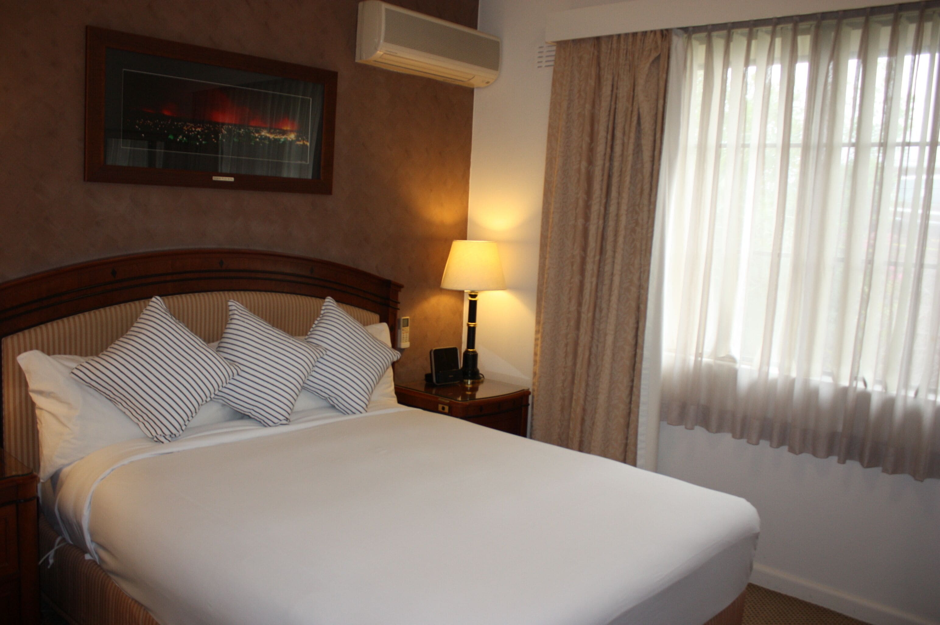 Habitación de hotel con una cama grande, una mesita de noche con lámpara, un cuadro enmarcado en la pared y una ventana con cortinas.