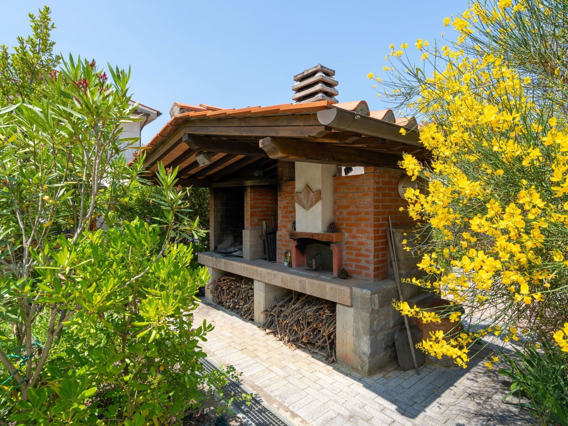 A rustic outdoor fireplace with a brick chimney, surrounded by greenery and yellow flowers.