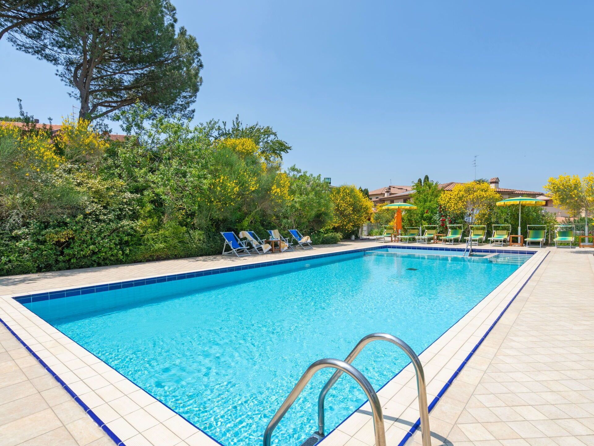 Ein rechteckiger Swimmingpool mit klarem, blauem Wasser, umgeben von einer gefliesten Liegefläche. Am Poolrand befinden sich Liegestühle und Sonnenschirme.