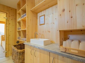 Een houten interieur met een witte wastafel, planken met handdoeken, een rieten mand en een ingelijst certificaat aan de muur.