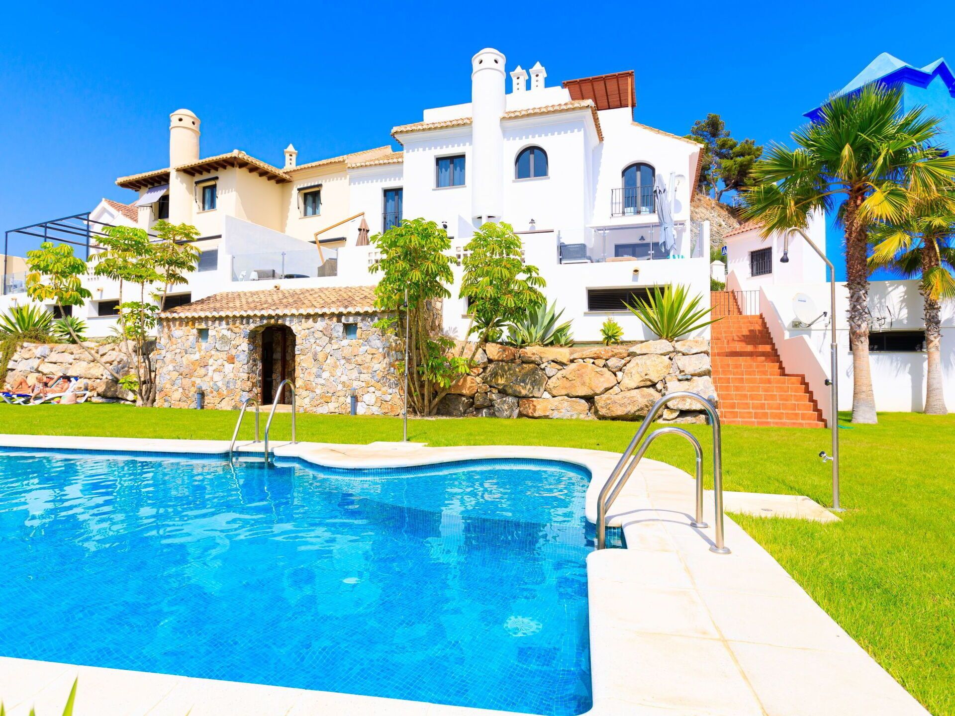 A swimming pool with a stone wall and a staircase leading to a white building with multiple balconies.