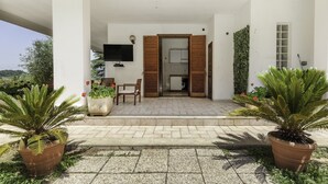 A covered outdoor area with a seating arrangement, a television mounted on the wall, and a door leading to a kitchen. - La Villa - Luxury Home (Configni)