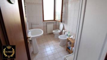 Un baño con lavamanos pedestal, dos inodoros, una ventana y un radiador.