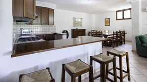 A kitchen with a central island, wooden cabinets, a gas stove, and a dining area with chairs and a table. - La Villa - Luxury Home (Configni)