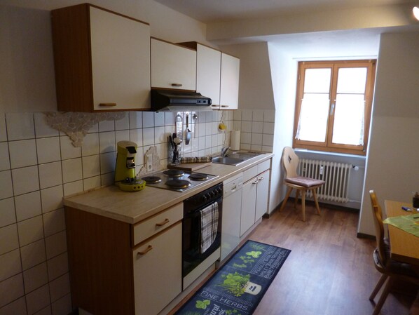 A compact kitchen with white cabinets, a stove, and a sink. There's a window with a view of the outside, and a radiator under the window.