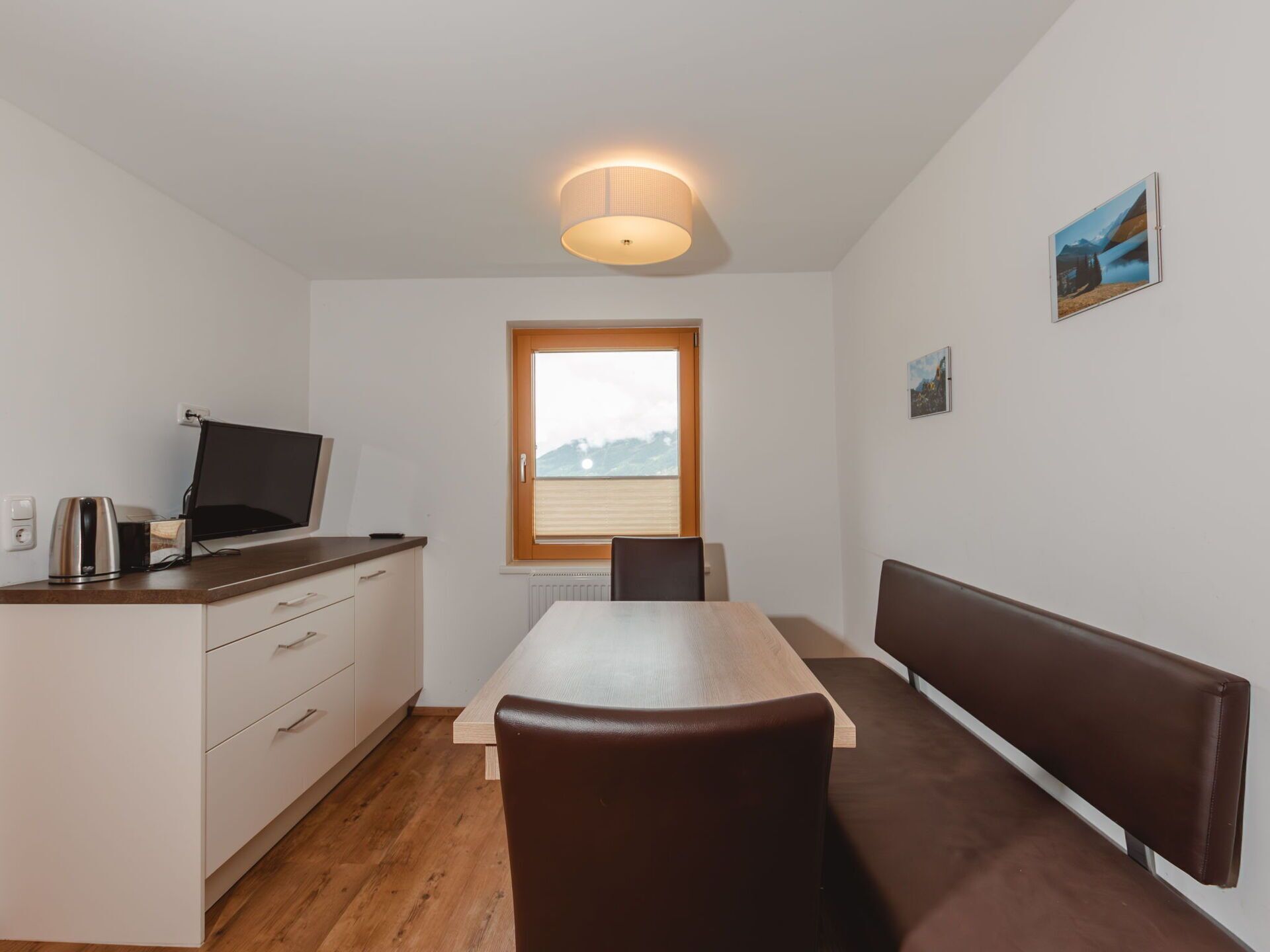 Eine kleine, moderne Küche mit Holzarbeitsplatte, einem Wasserkocher und einem Flachbildfernseher. Ein Fenster bietet Blick auf einen Berg und ein Bild hängt an der Wand.