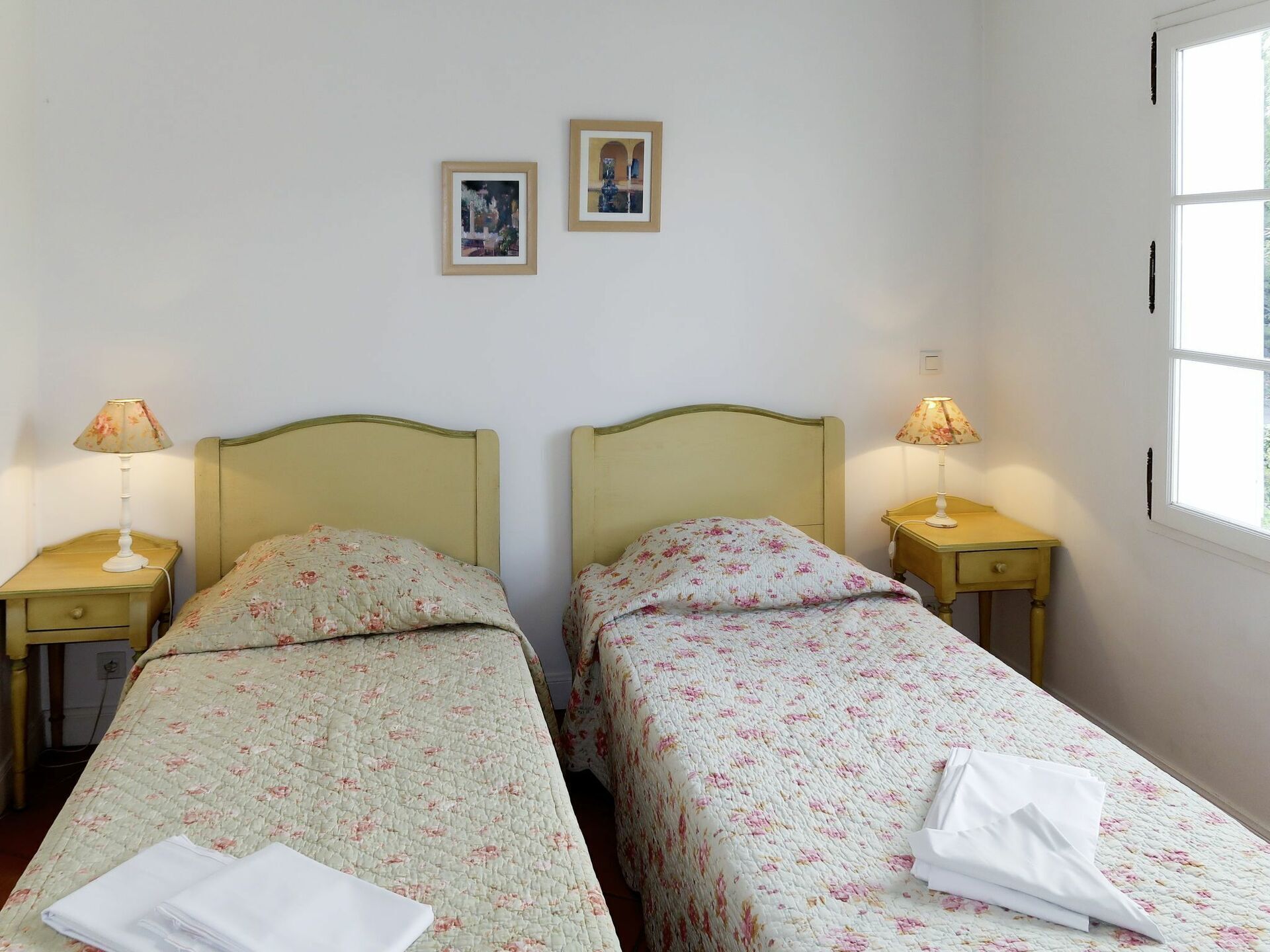 A twin room with two single beds, each with a floral bedspread and a matching nightstand with a lamp. There are two framed pictures on the wall.