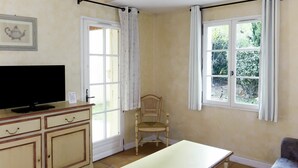 Una habitación con televisión sobre un mueble, una silla de madera, una mesa y dos ventanas con cortinas.