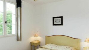 A bedroom with a bed, a nightstand with a lamp, a window with curtains, and a framed picture on the wall. - Lovely apartment in villa for 4 guests with A/C, pool, WIFI, TV, patio and pets allowed (Les Saquetons)